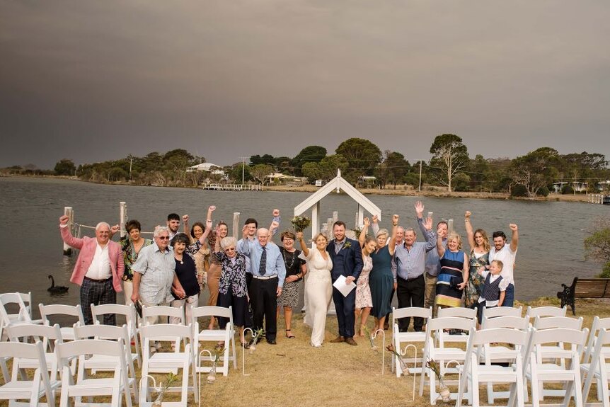 20 guests surround a bride and groom beside a lake a cheer with their arms in the air.
