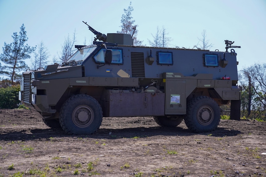 A grey-brown military vehicle is parked on dirt ground.