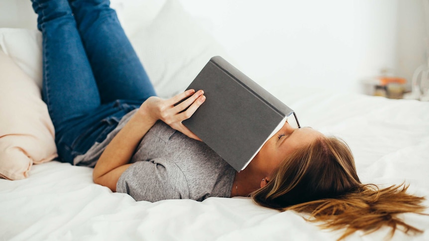Woman lying on bed reading book for a story about how reading erotica can unlock sex drive.