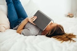 Woman lying on bed reading book for a story about how reading erotica can unlock sex drive.