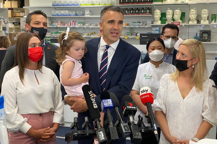 A man holding a small girl with other people standing around wearing face masks at a pharmacy