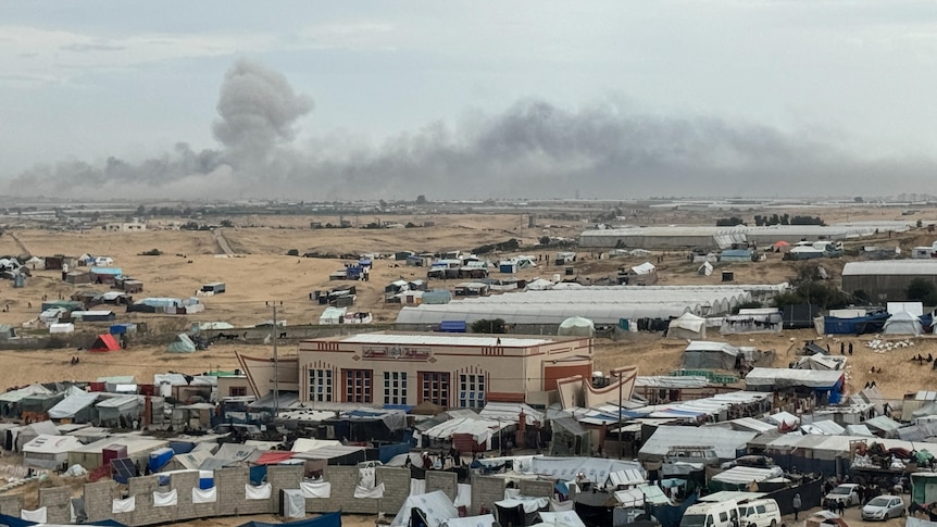 A ramshackle tent city full of tents and vehicles sits in the foreground as smoke rises from a city in the distance.