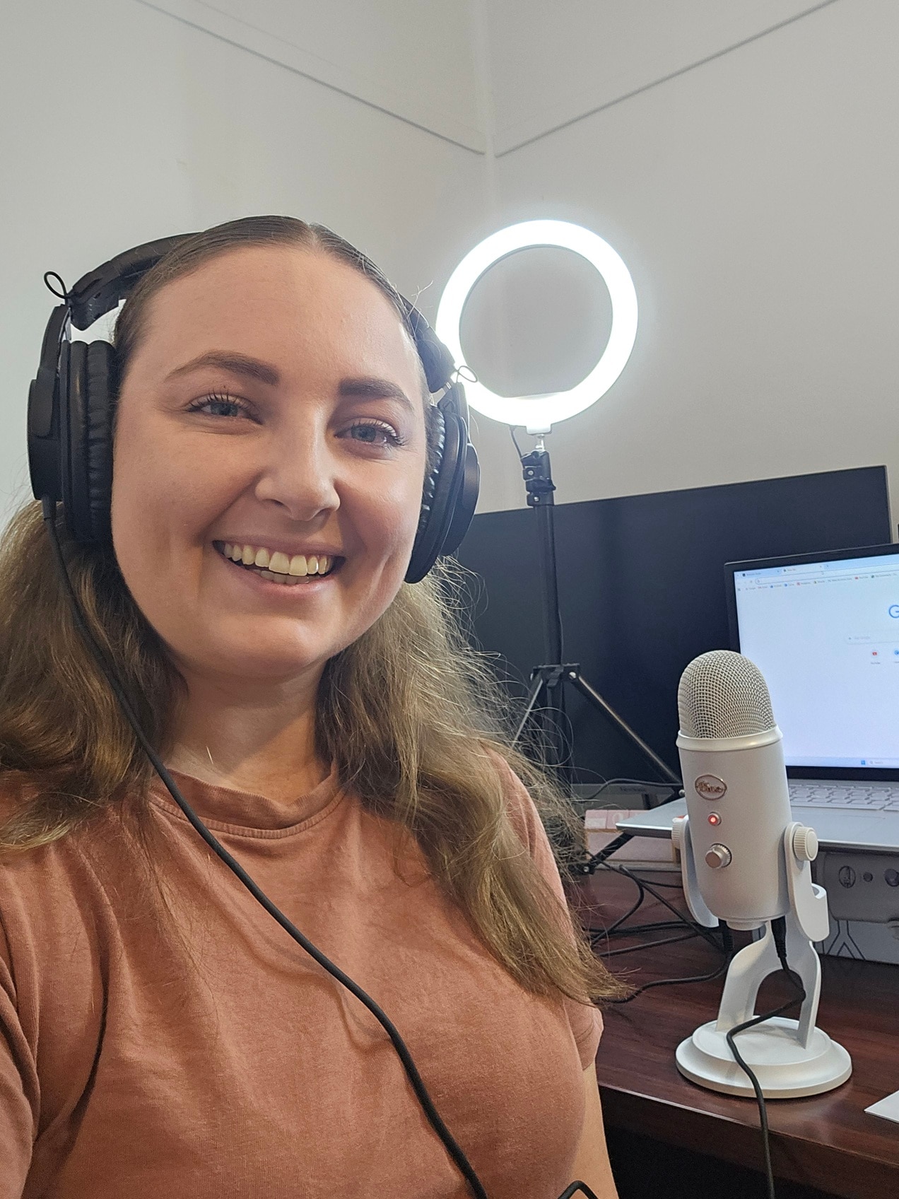 A woman in front of computer and microphone, wearing headphones, with a ring light in the background.