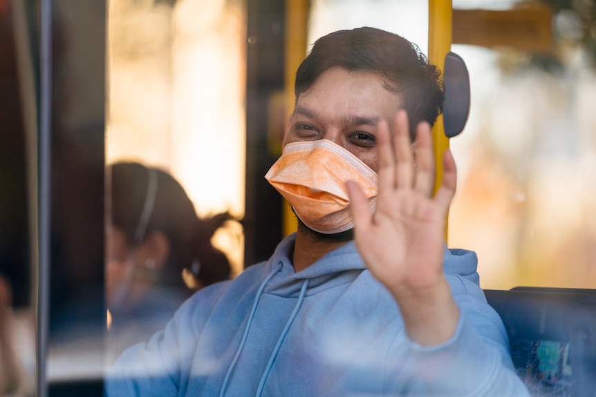 A man wearing a face mask waves as he looks out of the window of a bus.