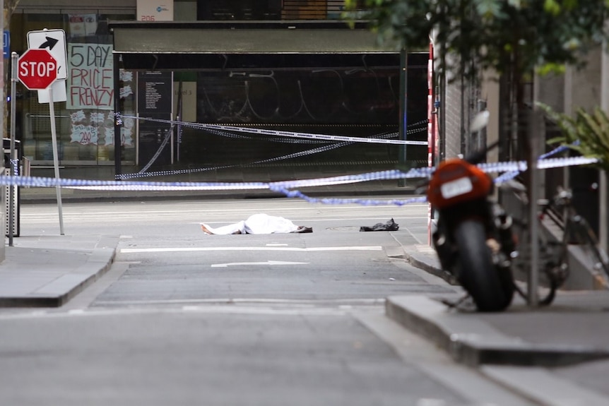 Police tape surrounds a body, covered by white material, lying on road.