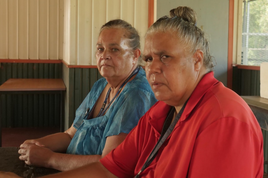 Two Aboriginal women sit in a school room, looking at the camera. 