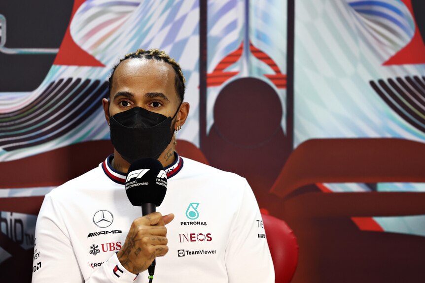A mask-wearing F1 driver holds a microphone at a press conference.