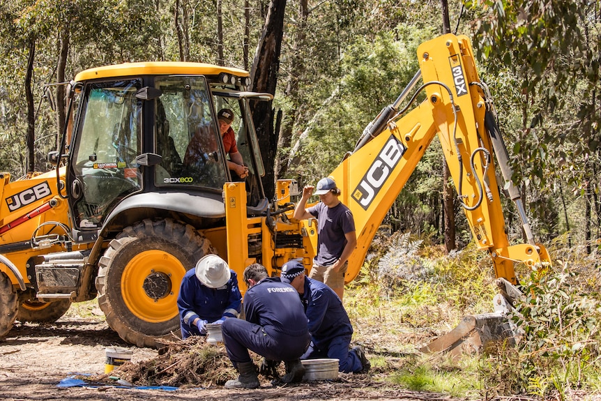 Three police on ground in front of pile of dirt while yellow digger and two men watch from behind.