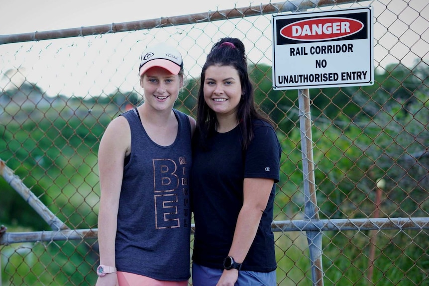Two girls stand next to each other and look at the camera. Behind them is a fence which has a danger no entry sign