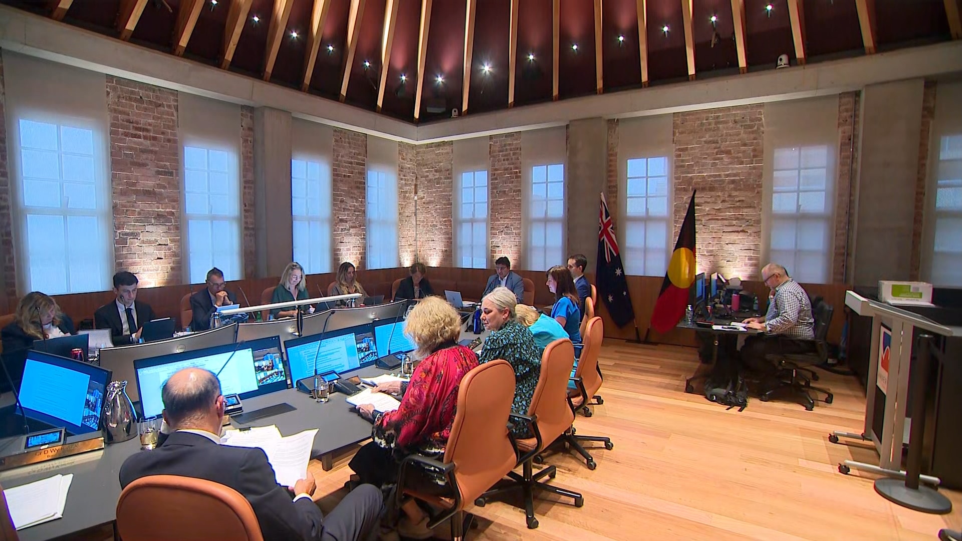 Una gran mesa de reuniones alargada donde se sientan los concejales de la ciudad en un edificio de techos altos.