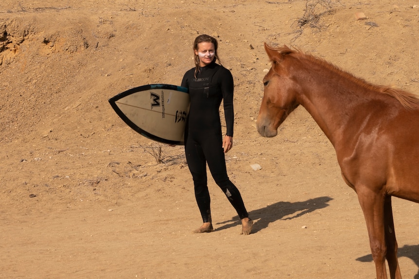 A woman holds a surfboard and looks at a horse standing nearby.