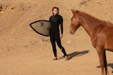 A woman holds a surfboard and looks at a horse standing nearby.
