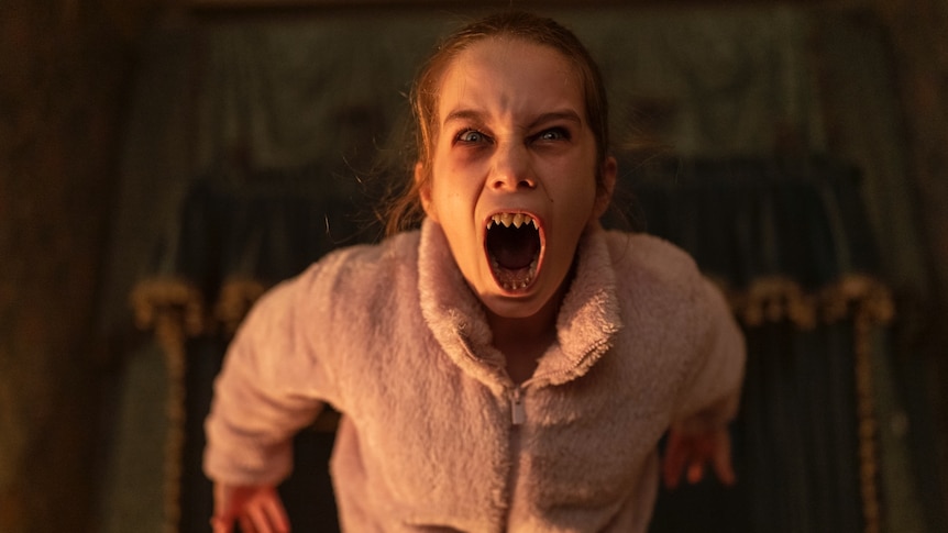 A young girl in a pink jacket with fake fangs screams at the camera