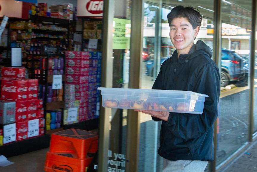 Smiling sixteen-year-old boy carries a box of freshly made doughnuts through supermarket doors to deliver