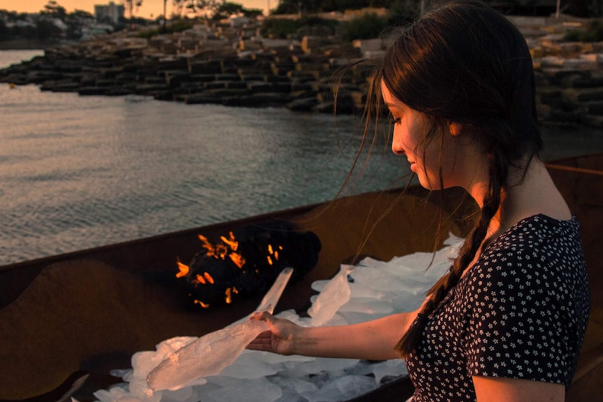 Young woman placing ice fish sculpture onto pile next to fire.