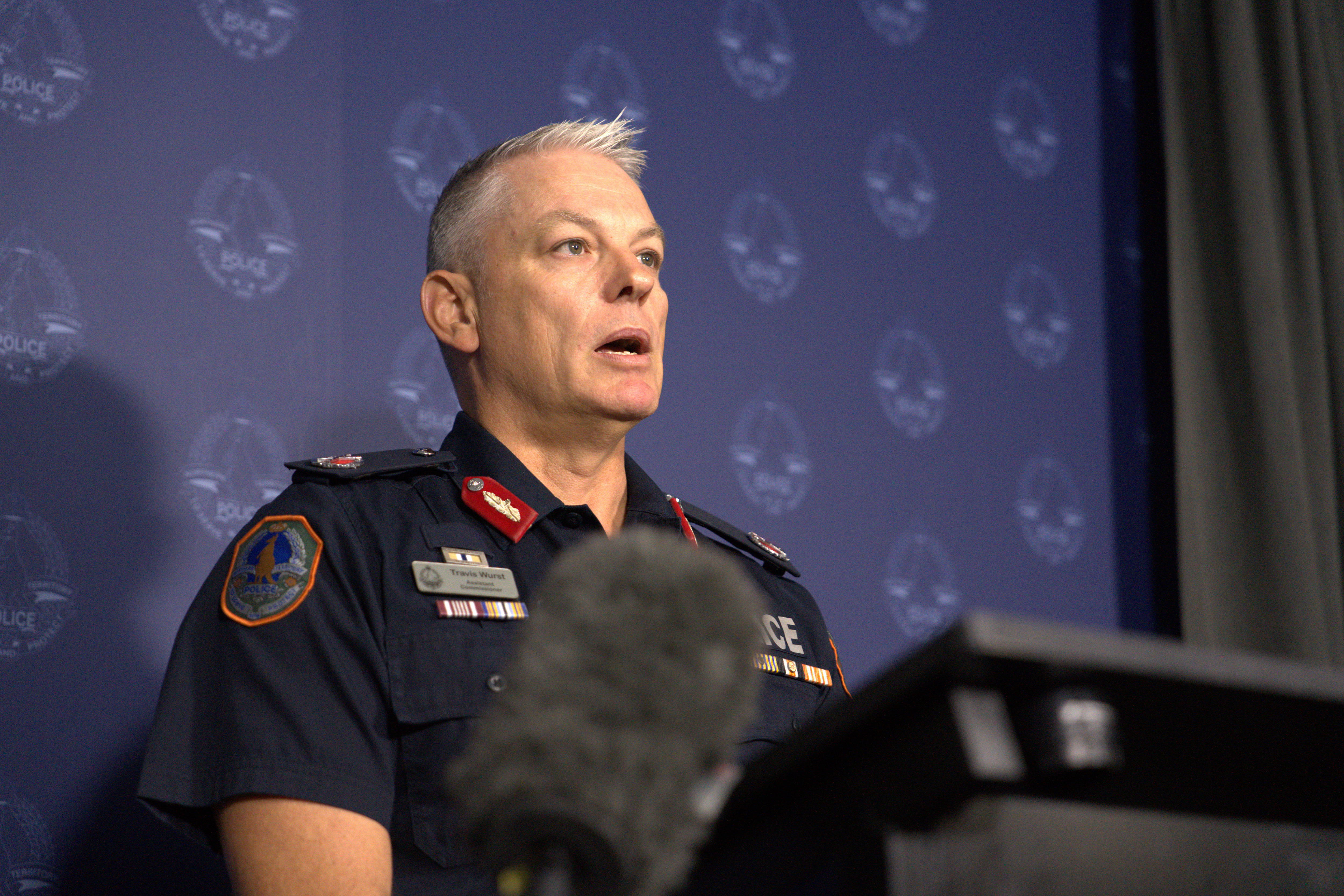 Un hombre de cabello claro con uniforme de la policía del NT y hablando por micrófonos de noticias.
