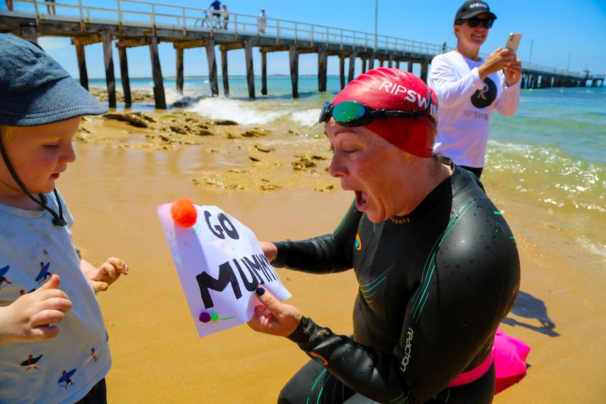 a woman in a wetsuit is excited to be given a sign that says "go mummy" from her child.