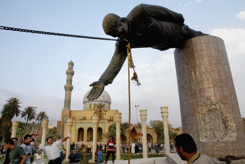 A large statue of Saddam Hussein is pulled from its plinth by a chain as people watch and hurl things 