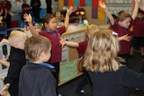 Primary school students stand in a classroom with their eyes closed and arms stretched out, practising mindfulness.