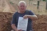 Mr Glew sits on a mound of sand and dirt, holding a piece of paper, blocking the driveway of his regional property.