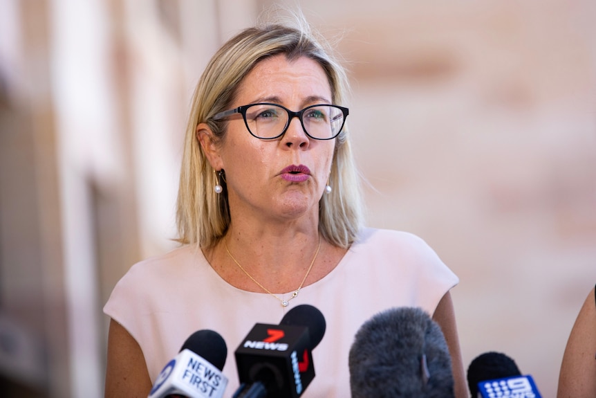 A woman with blonde hair and glasses speaking towards the right of frame.