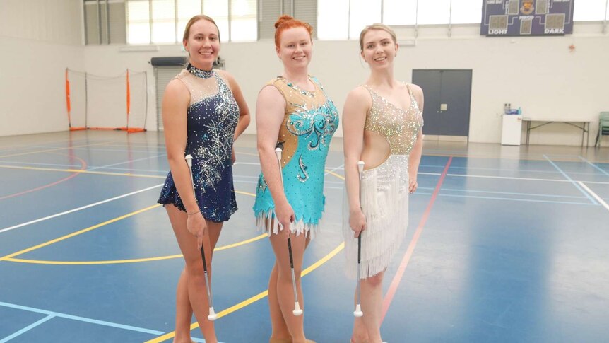 Three girls stand in a line wearing sparkly dance costumes with batons in their hand.