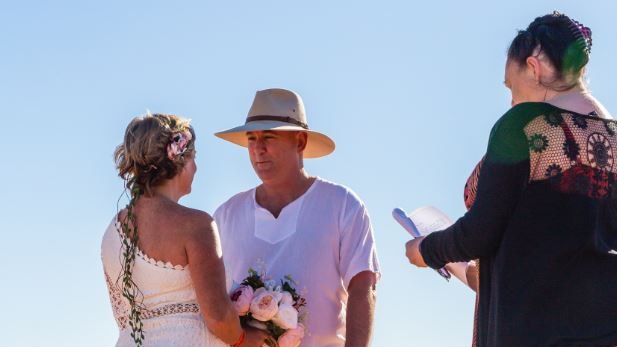A celebrant marries a couple who are standing on a hill with cars and tents in the background.