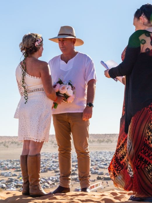 A celebrant marries a couple who are standing on a hill with cars and tents in the background.