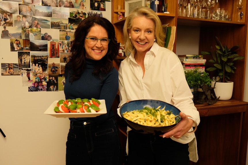 Two women holding bowls of food standing against a wall with family photos on it, smiling at the camera.