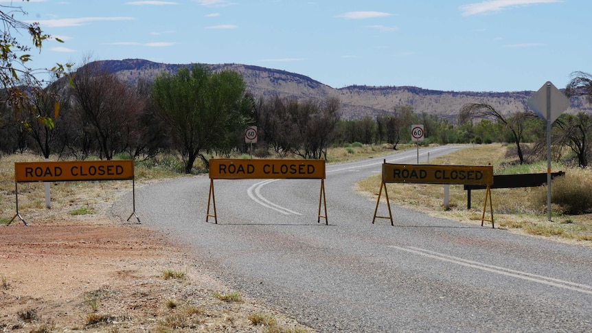 Three signs saying 'ROAD CLOSED' block a road leading to some ranges near Alice Springs.