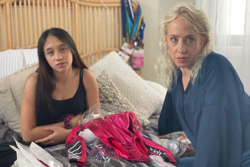 Stieve and Trinity De Lance sit on a bed surrounded by gymnastics costumes, looking sad and angry.