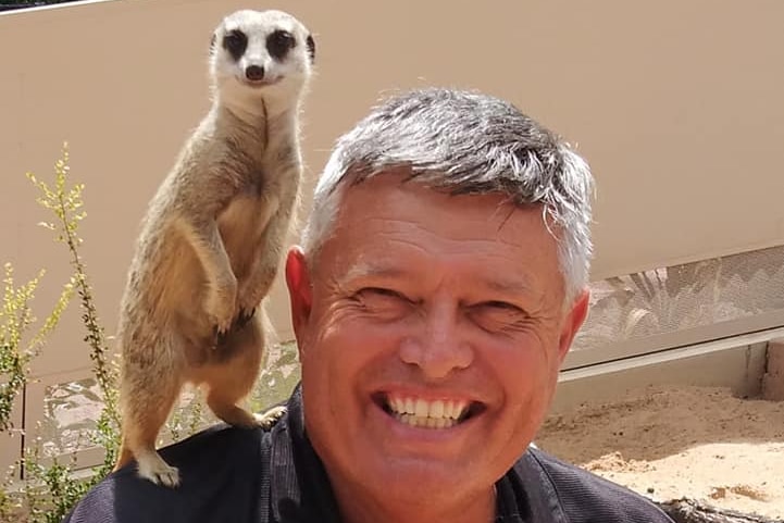 A man with a dark polo shirt. There is a meerkat on his shoulder.