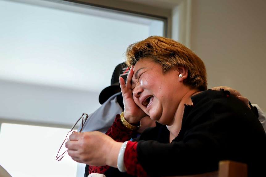 A woman with light-dyed hair cries, holding her head in one hand, glasses in another, as a person comforts her.