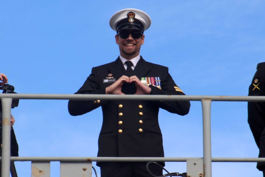 Man in full navy uniform makes a heart sign with his hands on the deck of navy ship.