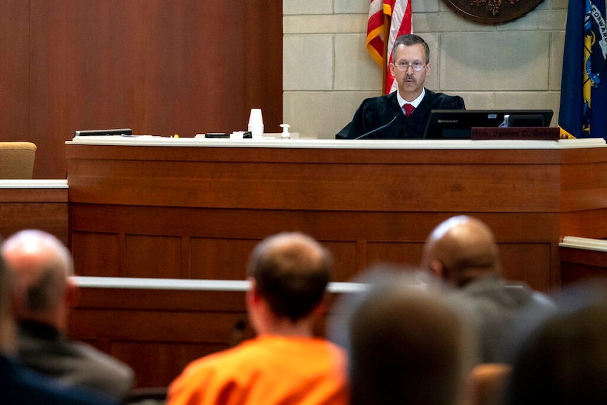 A judge wearing black court robes sits at a judicial bench, with a man in an orange prison uniform before him.
