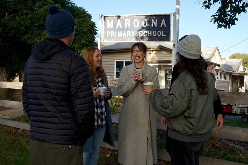 A woman holds a cup of coffee smiling standing with three other people. All are wearing winter gear and standing outside school
