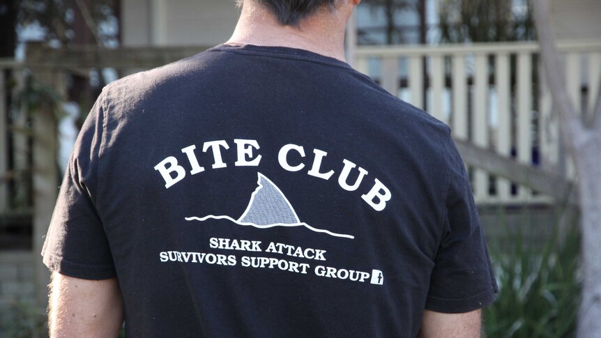 The back of a man wearing a tshirt. The back of the tshirt says "Bite Club: Shark Attack Survivors Support Group".