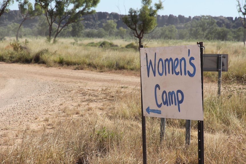 A sign saying "women's camp" on the side of a dirt road with grassland, trees and rocky mountains in the background.