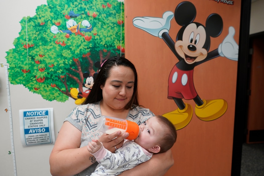 a woman feeds her daughter with a bottle in front of a door featuring a picture of Mickey Mouse