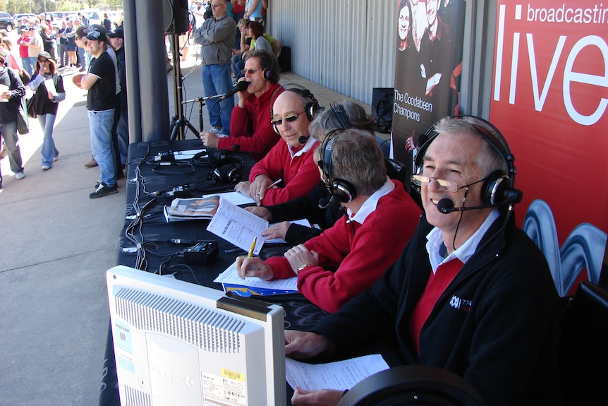 A row of men wearing red and black sit at a bench commentating outside at a football ground, with attendees in background