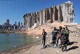 A group of emergency workers stand at a waterfront in front of a large crumbling concrete silo.