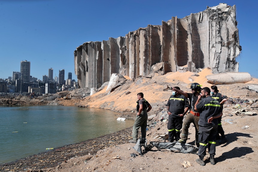 A group of emergency workers stand at a waterfront in front of a large crumbling concrete silo.