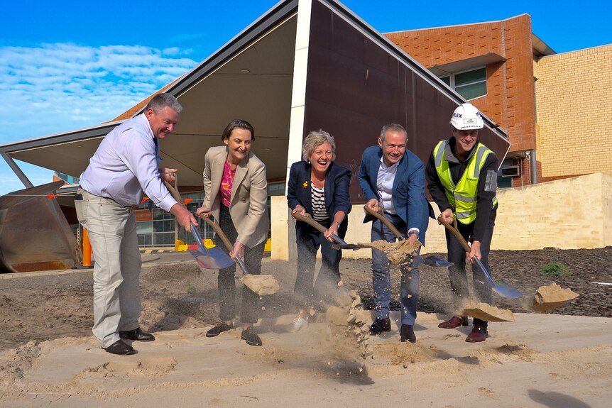 Two men and two women, three in formal wear and one in high-vis, shovel dirt outside a large building.