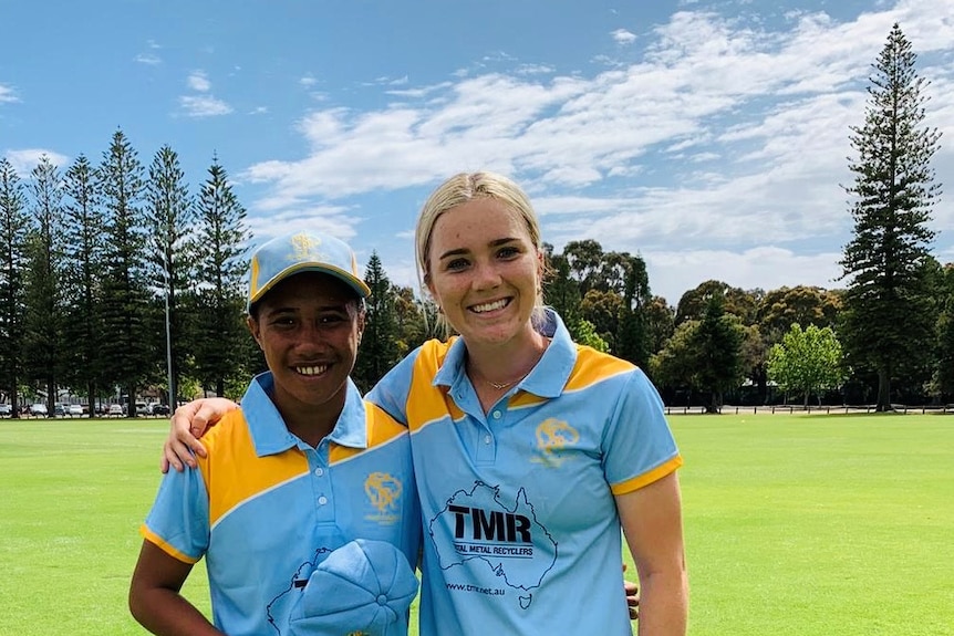 Two female cricketers, side by side, with their arms around each other. One has dark hair under a team cap, the other is blonde.