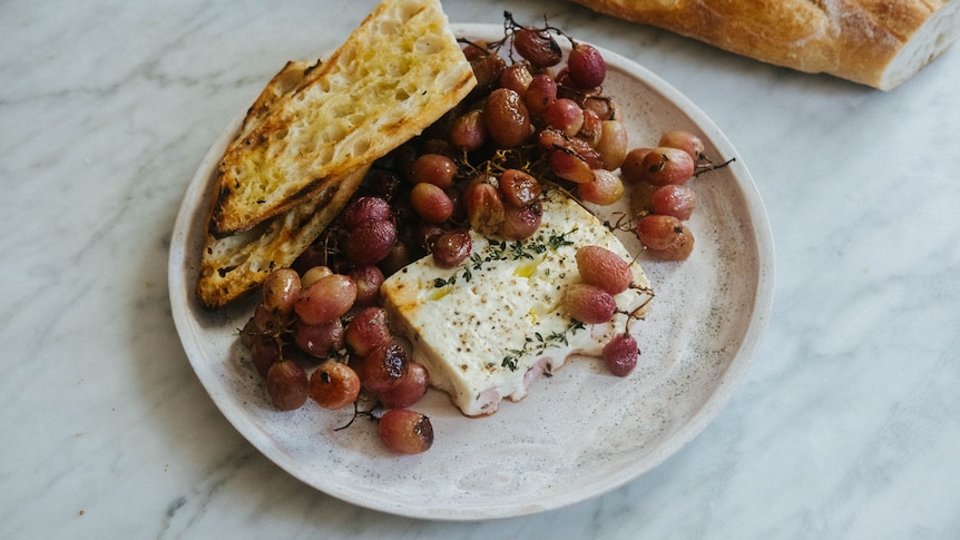 A block of feta cheese and red grapes, roasted on a tray with thyme to create a simple meal when served with a baguette