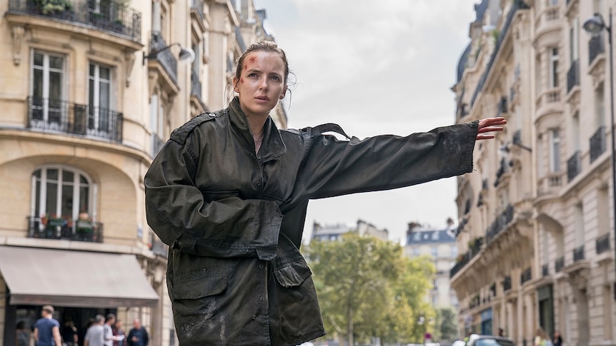 Actor Jodie Comer playing the character Villianelle in Killing Eve, she is hailing a taxi in a trenchcoat in a european street