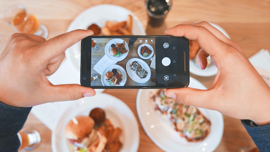 Two hands hold a mobile phone out over a meal on a table, taking a photo of the food