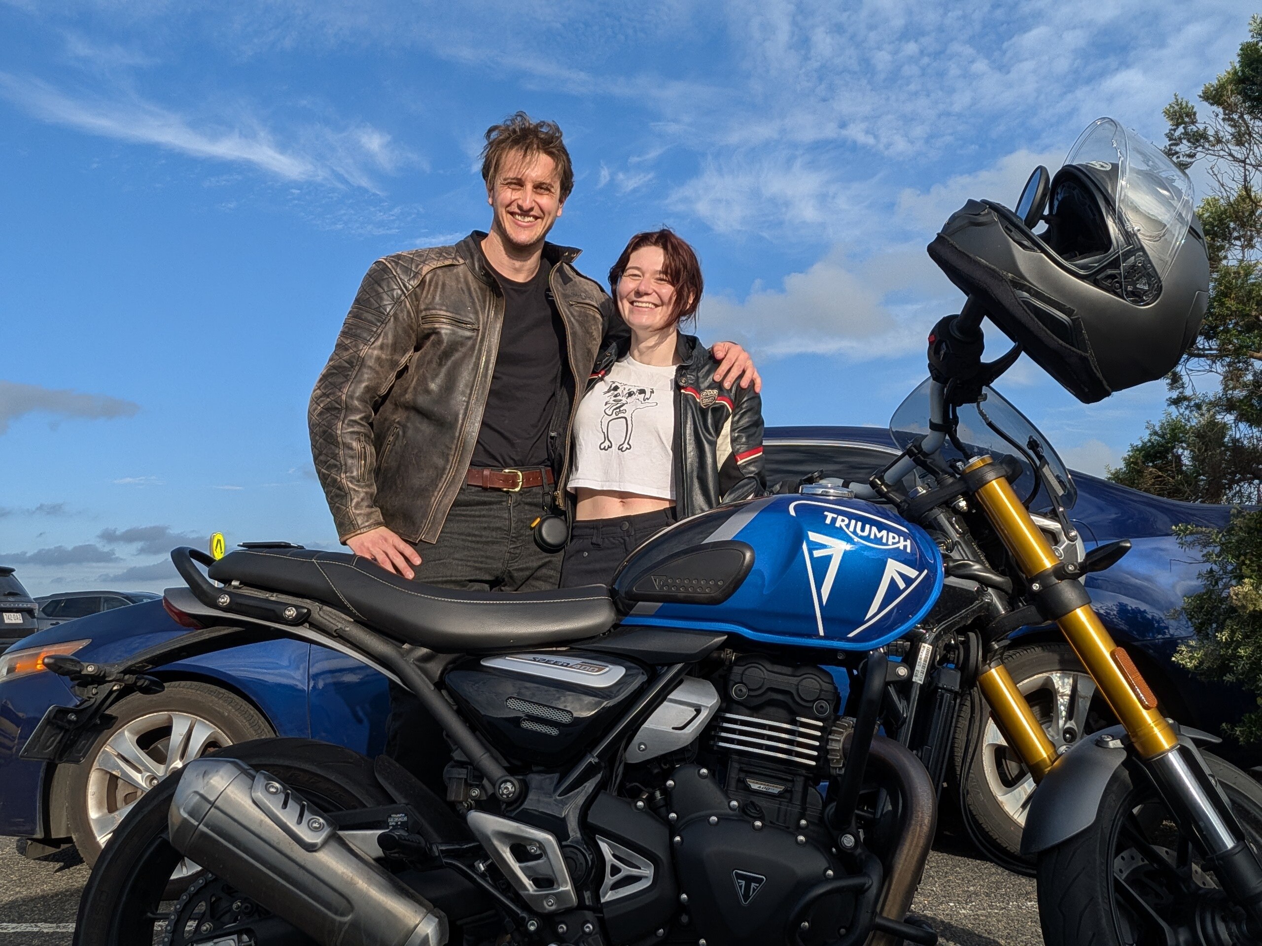 Una pareja posa detrás de una motocicleta en un día soleado.