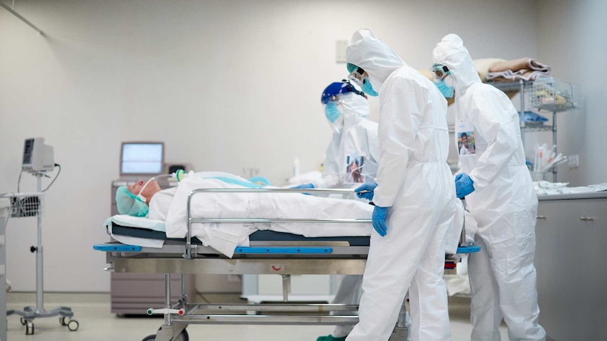 Healthcare workers in full PPE pushing a patient in a hospital bed.