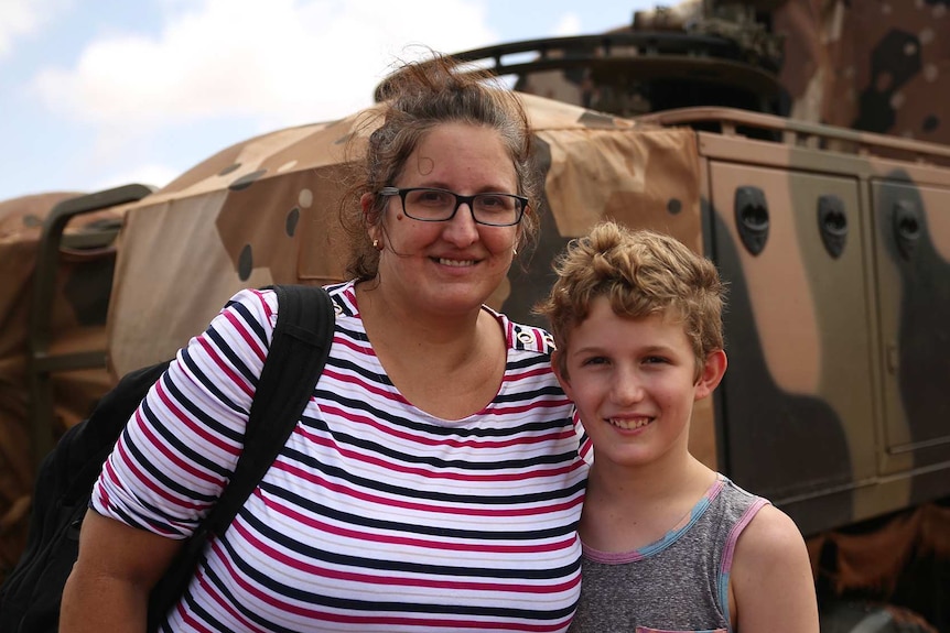 A woman and her son stand arm in arm in front of an army truck.